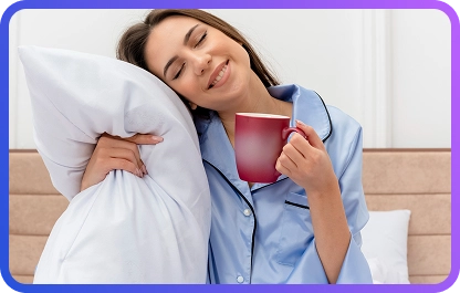 Een ontspannen vrouw in een blauwe pyjama, met een kussen en een rode mok, geniet van een rustige ochtend in een zachte, gezellige slaapkamer.